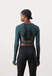 Top scurt cu mâneci lungi de culoare teal închis, cu spatele decupat și logo-ul "NIKE PRO" cu finisaj reflectorizant, purtat cu leggings negri cu talie înaltă.