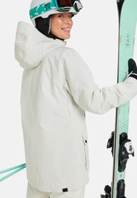 Weiße wasserdichte Skijacke mit Kapuze, strukturiertem Stoff und Reißverschlusstaschen, kombiniert mit mintfarbenen Skiern und schwarzen Bindungen.