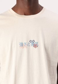 Beige pamut póló színes grafikai nyomattal, japán szöveggel és virágmintával a mellrészen. Klasszikus kerek nyakú stílus.