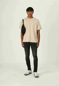 Jovem com cabelo preto em corte tipo tigela, vestindo t-shirt bege, jeans skinny pretos rasgados, ténis brancos e bolsa atravessada preta, contra fundo liso.