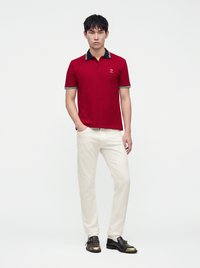 Jeune homme debout portant un polo rouge, un pantalon blanc et des chaussures noires avec des accents dorés sur un fond blanc uni.