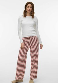 Femme debout portant une chemise blanche à manches longues, un pantalon large à rayures rouges et blanches, et des chaussures beige sur un fond uni.