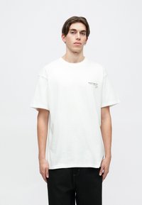 Biała bawełniana koszulka z krótkim rękawem, okrągłym dekoltem i małym czarnym logo na lewej piersi. Model nosi czarne spodnie.