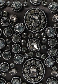 Czarna tkanina ozdobiona dużymi i małymi szarymi oraz przezroczystymi kamieniami szlachetnymi, w układzie okrągłym i owalnym, podkreślająca fakturę i blask.