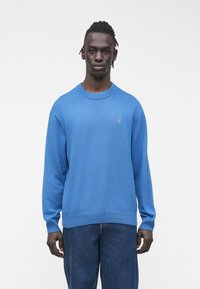 Moški stoji obrnjeno naprej, oblečen v modro majico z dolgimi rokavi in temno modre kavbojke na enostavni beli podlagi.