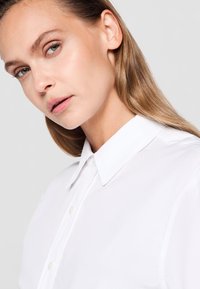 O femeie cu piele deschisă și păr blond drept poartă o cămașă albă cu guler, uitându-se ușor lateral, pe un fundal deschis gri uniform.