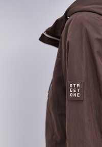 Brun jakkeærme med en firkantet "STREET ONE" logo-patch syet nær den øverste del af armen, vist mod en ensartet lys grå baggrund.