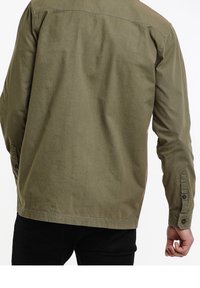 Camisa verde oliva de tejido suave, con cuello tipo botón, mangas largas, puños con botón y dobladillo recto.