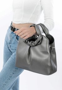 Borsa in pelle grigia con manico annodato, texture liscia e forma strutturata. Indossata con jeans leggeri blu e una maglietta bianca.