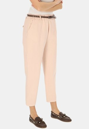 Kobieta w beżowych spodniach z wysokim stanem i zwężanych ku dołowi, z brązowym paskiem z ćwiekami i ciemnobrązowymi mokasynami, stojąca z założonymi ramionami.