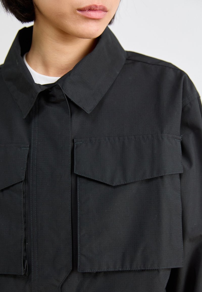 Veste noire à col pointu, avec deux poches poitrine et une fermeture à boutons sur le devant. Le tissu présente une texture subtile et des accents de coutures visibles.