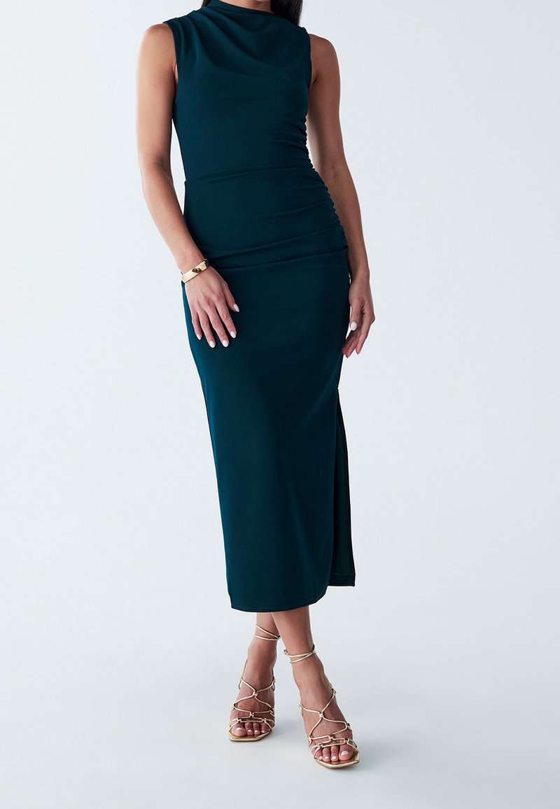 Femme portant une robe midi sans manches vert foncé avec une fente latérale et des sandales dorées à talons hauts à brides, debout sur fond blanc.