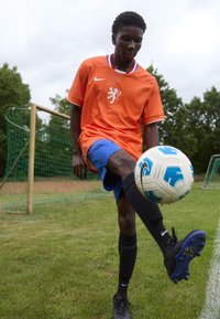 Oranje Nike voetbalshirt met een leeuw-logo, blauwe shorts, zwarte sokken en zwarte voetbalschoenen; speler controleert een wit en blauw voetbal.