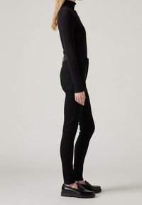 Femme debout en pull à col roulé noir, pantalon slim noir et mocassins noirs sur fond clair, vue de profil.