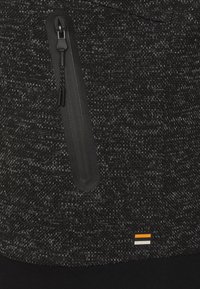 Sort tekstureret stof pullover med en sort lynlås-lomme, kontrasterende logo med orange og hvide accenter samt en ribbet sort bund.