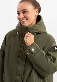 Giacca trapuntata verde oliva con colletto alto, hardware metallico e una patch a forma di cuore sulla manica. Presenta una finitura testurizzata.