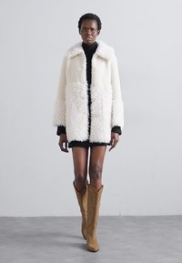 Witte fluweelachtige jas met een kraag, grote manchetten en vijf knopen, gecombineerd met een zwarte coltrui en een kort zwart jurkje. Bruine cowboylaarzen.