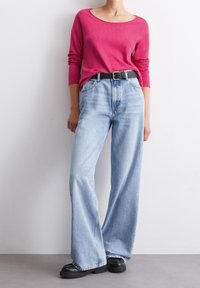 Långärmad rosa topp i kombination med ljusblå vida jeans, accentuerad med ett svart bälte och kraftiga svarta skor. Slät tyg och avslappnad passform.