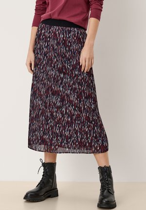 Vrouw draagt een geplooide midi-rok met een abstract patroon in rood, blauw en wit, gecombineerd met zwarte veterlaarsjes en een bordeauxrode top.