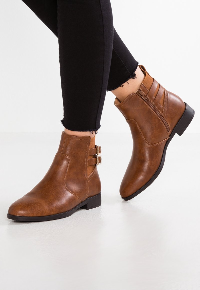 Bottines en cuir marron avec fermeture éclair latérale et doubles brides à boucle, portées avec un pantalon noir court à ourlets effilochés sur un sol blanc.