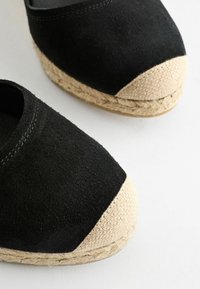 Espadrilles noires avec bouts tressés beiges et semelles en jute tressé sur fond blanc.