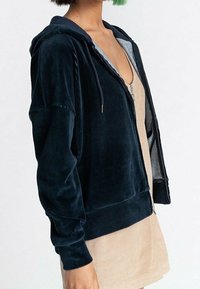 Navy corduroy zip-up hoodie with a grey-lined hood. Features a relaxed fit, ribbed cuffs, and a smooth texture. Paired with a beige dress.