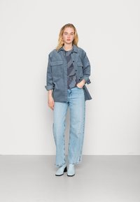 Jaqueta de denim em azul claro com dois bolsos no peito, sobre uma blusa escura de padrão. Jeans de perna larga em azul claro e botas de tornozelo combinando.