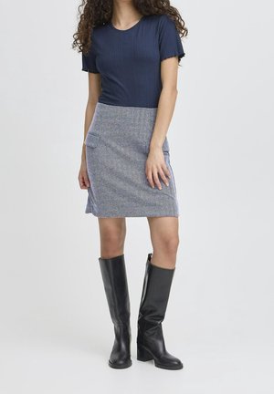 Kobieta ubrana w granatowy prążkowany top z krótkim rękawem, szarą mini spódniczkę w wzory z kieszeniami oraz czarne, sięgające kolan botki na obcasie, stojąca na białym tle.