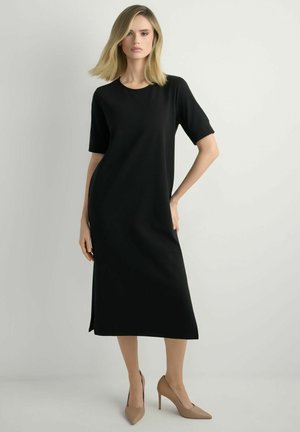 Blondynka stoi w czarnej sukience midi z krótkimi rękawami i beżowych szpilkach na tle jasnego, jednolitego tła.