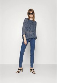 Mulher usando óculos de sol pretos, blusa azul de padrão solto, jeans azuis justos e sandálias pretas de salto, de pé contra uma parede branca.