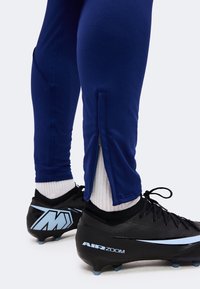Jambes en pantalon de sport bleu marine avec fermetures éclair aux chevilles, chaussettes blanches et chaussures de football noires avec « AIR ZOOM » et accents bleus.