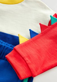 Farverig børnesweater med hvid overdel, bl�å nederste sektion, scallopede multicolore pigge og en rød ribbet manchet.