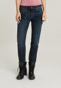 Dunkelblaue Slim-Fit-Jeans mit mittelhoher Taille, fünf Taschen und sichtbaren Nähten, kombiniert mit schwarzen Schnür-Stiefeletten.