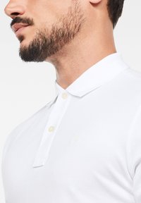 Vit polo-shirt med en texturerad tyg, tre knappar och en kort krage. Har en subtil logotyp broderad på bröstet i matchande vitt.