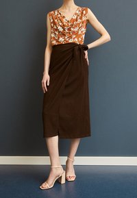 Brun omslagkjol tillsammans med en orange blommig topp med krage, som har mjuka texturer och knälång längd. Ljust beige blockklack-sandaler.