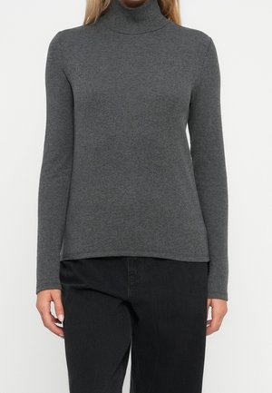 Kobieta ubrana w dopasowany szary grafitowy golf z długim rękawem i czarne spodnie, stojąca na tle jednolitego białego tła.