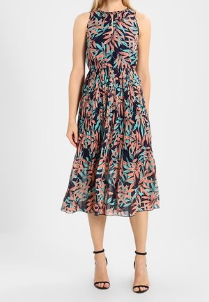 Robe midi sans manches avec base bleu marine et imprimé de feuilles corail et bleu sarcelle, taille froncée, portée avec des talons noirs à bride à la cheville.