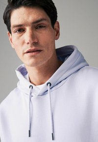 Helle lila Kapuzenjacke mit Kordelzug und silbernen Ösen, mit einer glatten Textur und einem lockeren Schnitt, über einem weißen Shirt getragen.
