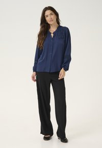 Blouse bleu marine à manches longues avec des boutons, détails en dentelle florale sur les épaules, poignets froncés, assortie à un pantalon large noir.