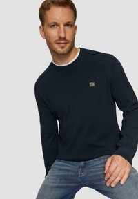 Marineblauwe gebreide trui met een ronde halslijn, geribde manchetten en een klein logopatch op de borst, in combinatie met lichtblauwe jeans.