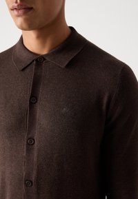 Gros plan d'une personne portant une chemise en tricot marron foncé à boutons avec col et un logo brodé discret sur la poitrine.