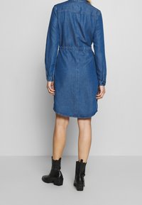 Kvinna som står vänd bort med en knälång denimklänning med långa ärmar och svarta ankelstövlar mot en enfärgad bakgrund.