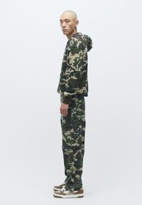 Kleita ar kamuflāžas rakstu, kas izgatavota no kokvilnas maisījuma auduma, ar brīvu piegriezumu, garām piedurknēm un saskaņotām kravas kabatām, kopā ar sporta apaviem.