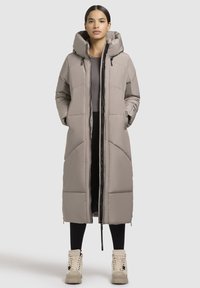 Dlhý prešívaný kabát svetlo béžovej farby, s kapucňou, predným zipsom a prešívanou textúrou. Skombinovaný s čiernymi legínami a béžovými vysokými teniskami.