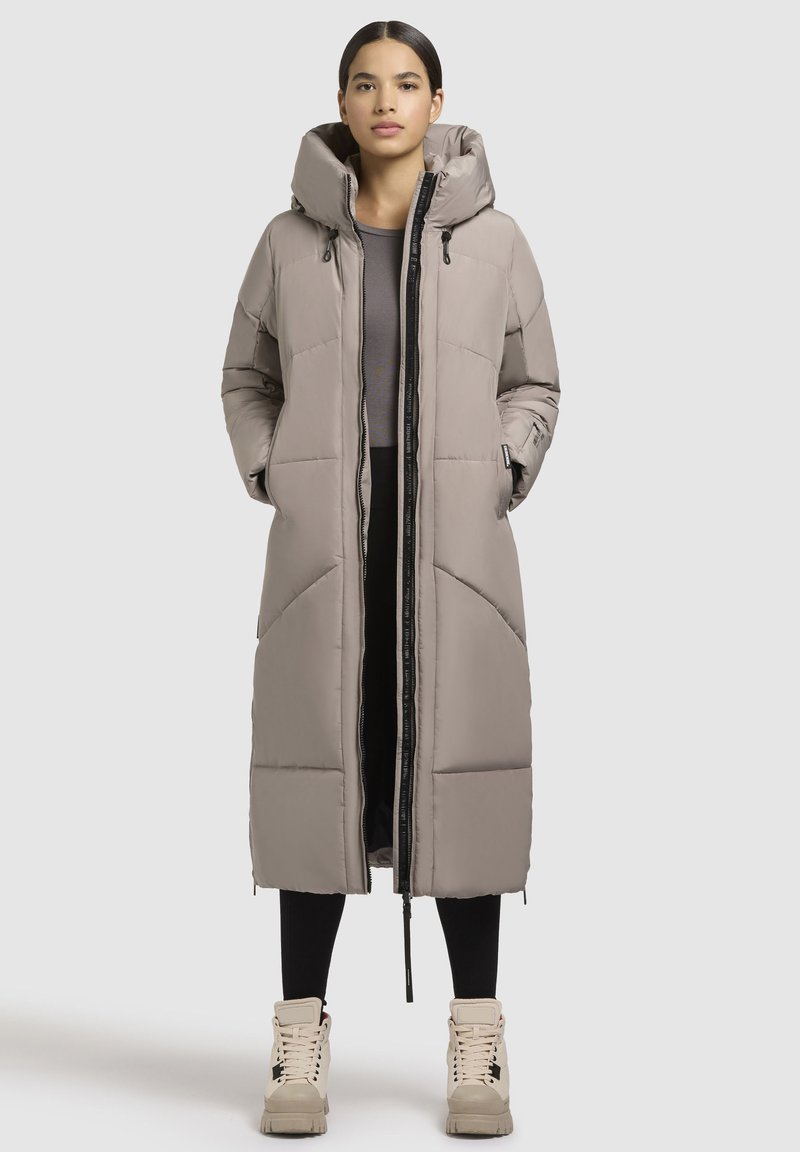 Ilga pūkinė striukė šviesiai béžinė, su kapuce, užtrauktuku priekyje ir iškloto tekstūros. Su juodomis leginsinėmis kelnėmis ir béžiniais aukščiais sportbačiais.