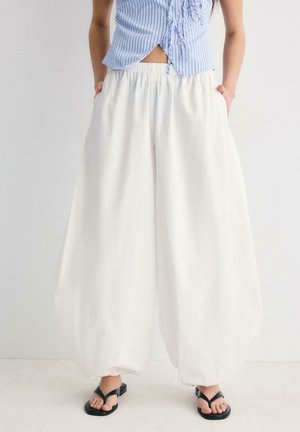 Kobieta ubrana w luźne białe spodnie typu balloon z elastycznym paskiem i czarnosandale, ręce w kieszeniach, stoi na tle jednolitego jasnego tła.
