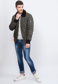 Olivgrüne Bomberjacke mit einem braunen Kunstfellkragen, Reißverschlusstasche am Ärmel, kombiniert mit hellblauen Jeans und weißen Sneakers.