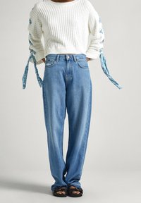 Suéter blanco con ribetes y lazos azules en las mangas, combinado con jeans de mezclilla de pierna ancha en un tono de azul claro y sandalias negras.