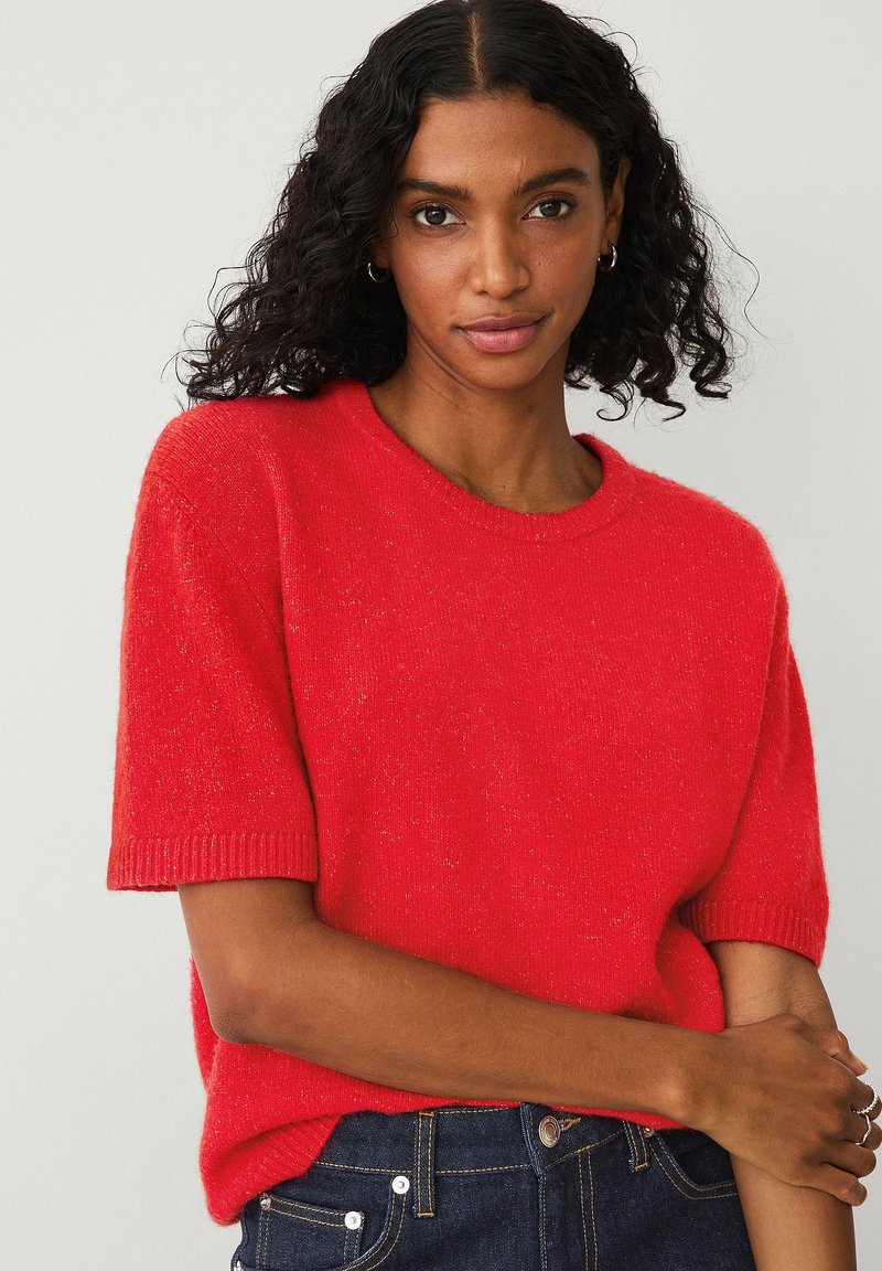 Rød kortærmet strikket sweater med rund hals, ribbede manchetter og en tekstureret finish, båret med mørkeblå jeans.