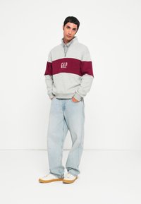 Gray sweatshirt with maroon stripe, "GAP" logo in white, half-zip collar. Paired with light blue jeans and cream shoes. Simple, casual fit.